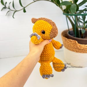 May include: A handmade, plush, yellow and brown crocheted horse. The horse has gray accents on its hooves and mane. The horse is being held in a person's hand. The text "TANATIcrochet by Tatiana Kucherovska" is visible in the image.