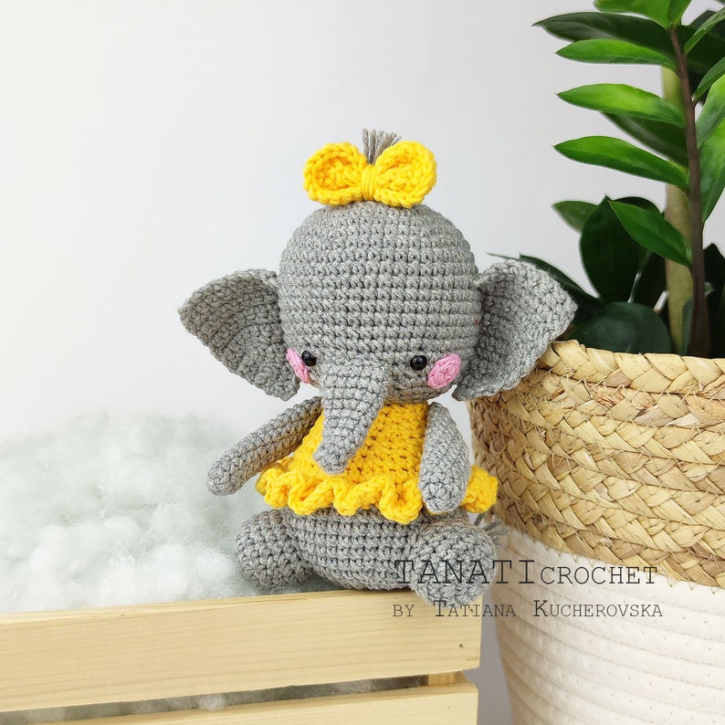Pode incluir: Um elefante de croch&ecirc; cinza vestindo um vestido de croch&ecirc; amarelo e um la&ccedil;o amarelo. O elefante est&aacute; sentado em uma caixa de madeira.