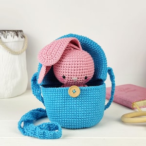May include: A crocheted blue bag with a pink crocheted bunny inside. The bag has a strap and a wooden button.