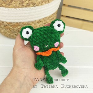 May include: A green crocheted frog with white eyes, pink cheeks, and an orange scarf. The frog is being held in a hand.