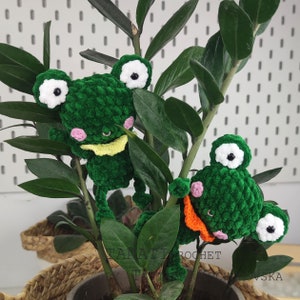 May include: Two green crocheted frog plushies with white eyes and pink cheeks. The frogs are sitting on a green plant with brown leaves.