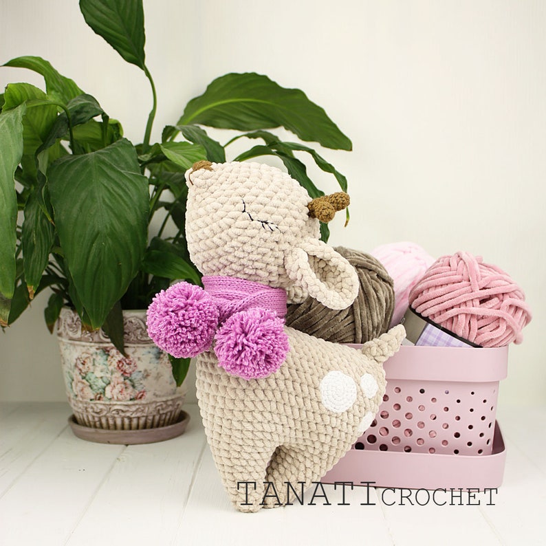 May include: A crocheted fawn plush toy with a pink scarf and pom poms. The fawn is standing on a white surface next to a pink basket with yarn.