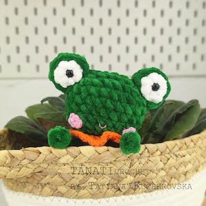 May include: Green crocheted frog amigurumi toy with white eyes and pink cheeks. The frog is wearing an orange scarf and is sitting in a woven basket.