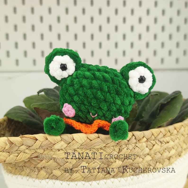 May include: Green crocheted frog amigurumi toy with white eyes and pink cheeks. The frog is wearing an orange scarf and is sitting in a woven basket.