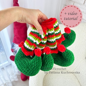 May include: Crocheted elf slippers in green, red, and white. The slippers have a green foot, red trim, and striped leg warmers. The image includes the text "+ video tutorial" and the ATlcrochet logo.