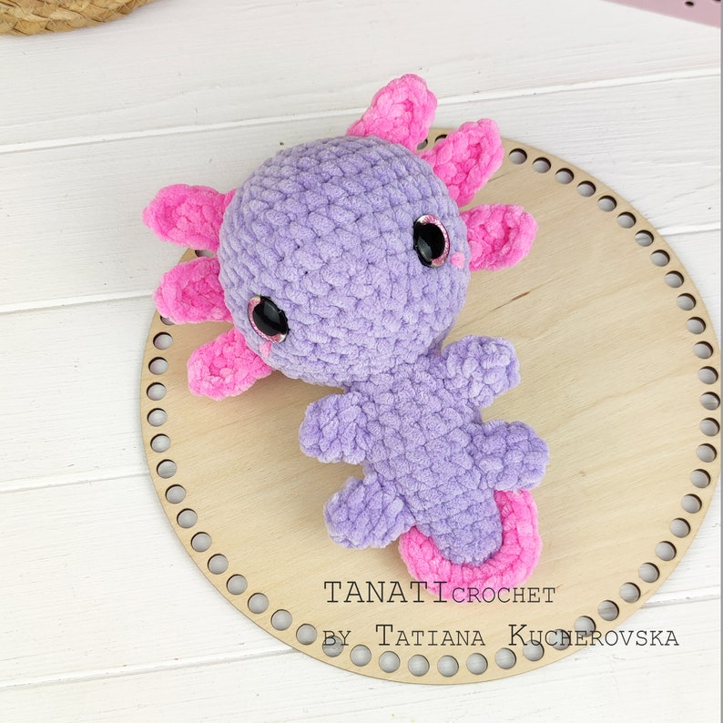 May include: A crocheted purple axolotl plush toy with pink accents and black eyes. The toy is sitting on a wooden circle with the text "TANATI CROCHET BY TATIANA KUCHEROVSKA" on it.