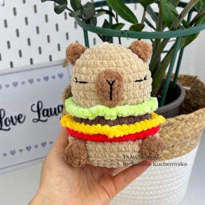 以下が含まれることがあります： ハンバーガーのデザインの、手作りのかぎ針編みラマぬいぐるみ。ラマはベージュで茶色のアクセントがあり、ハンバーガーは緑、黄色、赤の糸でできています。ぬいぐるみは手で持たれており、背景には植物と「Love Llama」と書かれたサインがあります。