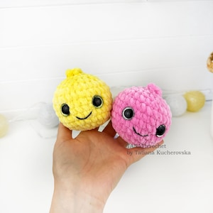 May include: Two round, plush, crocheted toys, one yellow and one pink, each featuring large black safety eyes and a stitched smiling mouth. The text TANATicrochet by Tatiana Kucherovska is visible in the lower right corner of the image.