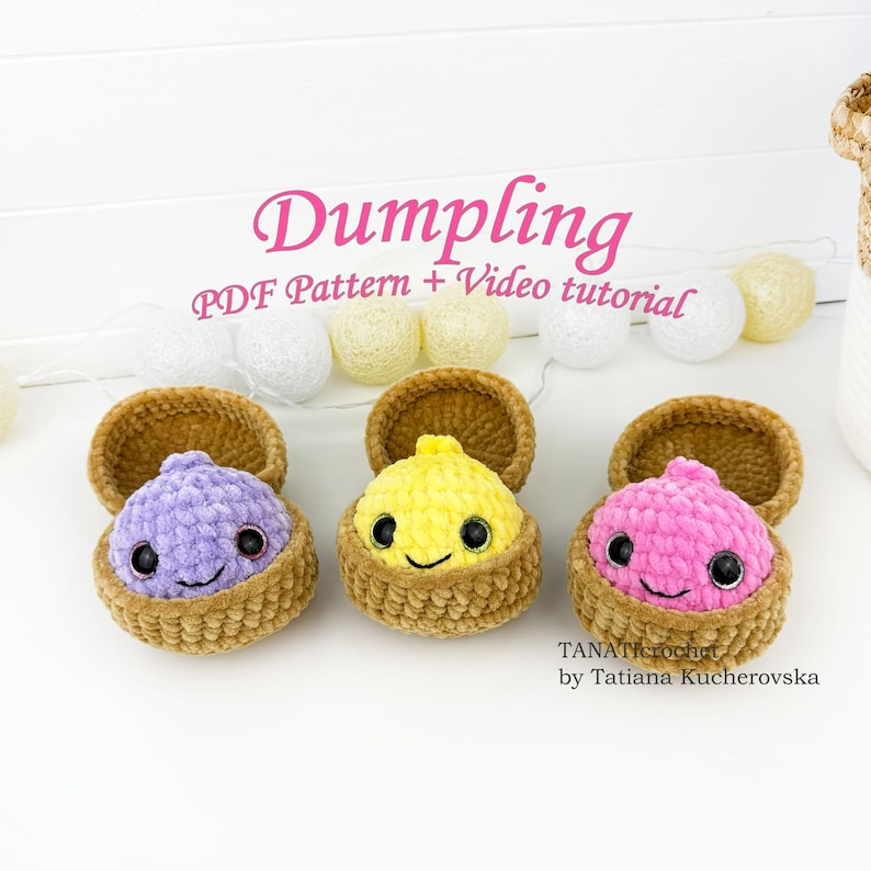 May include: Three crocheted plush dumplings in purple, yellow, and pink, each nestled in a brown textured container. The text reads Dumpling PDF Pattern + Video tutorial and TANAcrochet by Tatiana Kucherovska. The items are arranged in a row.