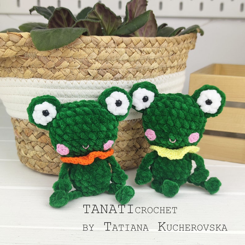 May include: Two green crocheted frog plushies with white eyes and pink cheeks. One frog has an orange scarf and the other has a yellow scarf. The frogs are sitting on a white surface. The text "TANATI CROCHET BY TATIANA KUCHEROVSKA" is below the frogs.