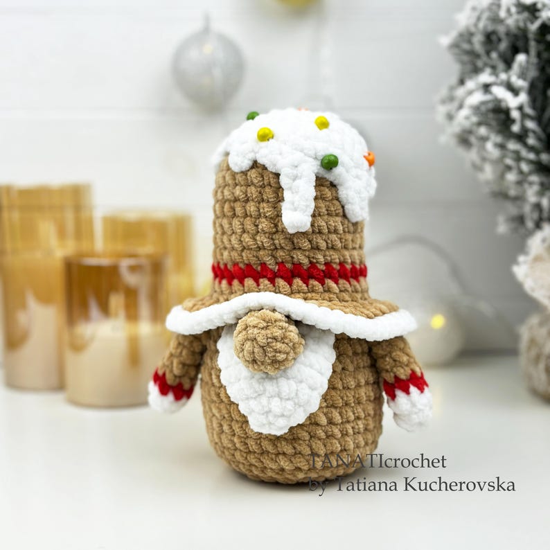 K&ouml;nnte beinhalten: Geh&auml;kelte Lebkuchenmann-Puppe mit wei&szlig;er Zuckerguss-Oberseite und bunten Streuseln. Die Puppe hat einen braunen K&ouml;rper, einen roten Streifen und wei&szlig;e Verzierungen. Arme und F&uuml;&szlig;e haben rot-wei&szlig;e Details. Eine festliche Weihnachtsdekoration.