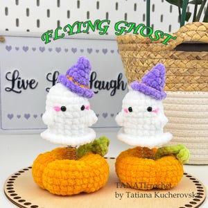 Puede incluir: Dos figuras de fantasmas de ganchillo hechas a mano con sombreros de bruja morados, mejillas rosas y ojos negros, sobre calabazas naranjas. Las calabazas se asientan sobre una base de madera. El texto "FLYING GHOST" y "Live Laugh" son visibles.