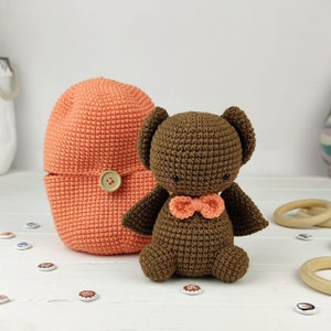 Puede incluir: Un juguete de murciélago de crochet marrón con una pajarita de color melocotón, sentado junto a un gorro de crochet de color melocotón con un botón.