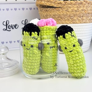 May include: A clear glass jar filled with three green, handmade, Frankenstein-themed crochet dolls. Each doll has black hair, gray arms, and a stitched face. One doll is outside the jar. The text "TANATlcrochet by Tatiana Kucherovska" is visible.