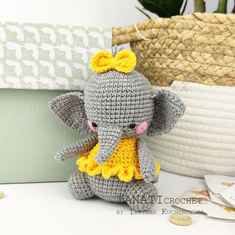 Pode incluir: Um elefante de croch&ecirc; cinza vestindo um vestido de croch&ecirc; amarelo com um la&ccedil;o amarelo na cabe&ccedil;a. O elefante est&aacute; sentado em uma superf&iacute;cie branca.