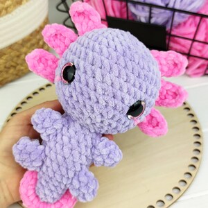 May include: A crocheted axolotl plush toy with pink fins and purple fur. The toy has large black eyes with glitter accents.