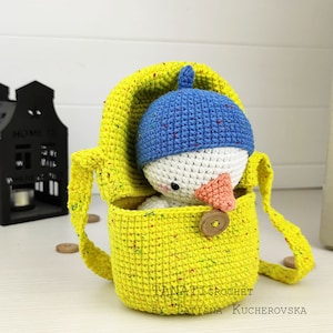 May include: A small crocheted snowman wearing a blue hat is inside a yellow crocheted bag with a strap. The bag has a wooden button closure. The text "TANATICROCHET BY TATIANA KUCHEROVSKA" is at the bottom of the image.