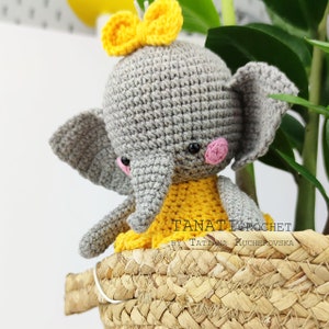 Pode incluir: Um elefante de croch&ecirc; cinza com um la&ccedil;o amarelo na cabe&ccedil;a. O elefante est&aacute; vestindo um vestido de croch&ecirc; amarelo e tem bochechas cor-de-rosa. O elefante est&aacute; sentado em uma cesta de vime.