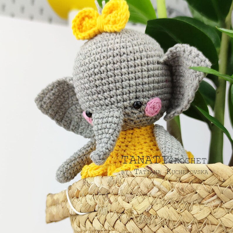 Pode incluir: Um elefante de croch&ecirc; cinza com um la&ccedil;o amarelo na cabe&ccedil;a. O elefante est&aacute; vestindo um vestido de croch&ecirc; amarelo e tem bochechas cor-de-rosa. O elefante est&aacute; sentado em uma cesta de vime.
