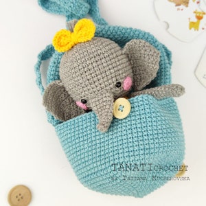Pode incluir: Um elefante de croch&ecirc; cinza com um la&ccedil;o amarelo est&aacute; sentado dentro de uma bolsa de croch&ecirc; azul com um bot&atilde;o amarelo. A bolsa tem uma al&ccedil;a e &eacute; feita com um ponto texturizado.