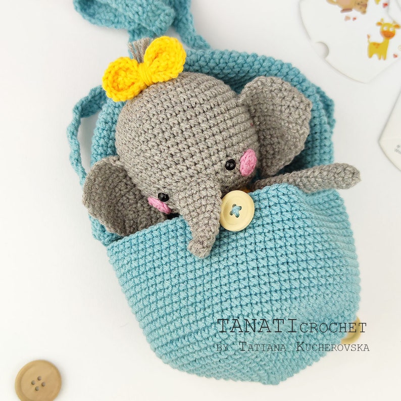 Pode incluir: Um elefante de croch&ecirc; cinza com um la&ccedil;o amarelo est&aacute; sentado dentro de uma bolsa de croch&ecirc; azul com um bot&atilde;o amarelo. A bolsa tem uma al&ccedil;a e &eacute; feita com um ponto texturizado.