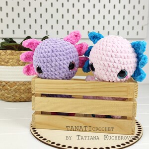 May include: Two crocheted axolotl plushies, one purple and one pink, with pink and blue accents, sitting in a wooden crate. The crate is on a wooden base with the text "TANATI CROCHET BY TATIANA KUCHEROVSKA" on it.