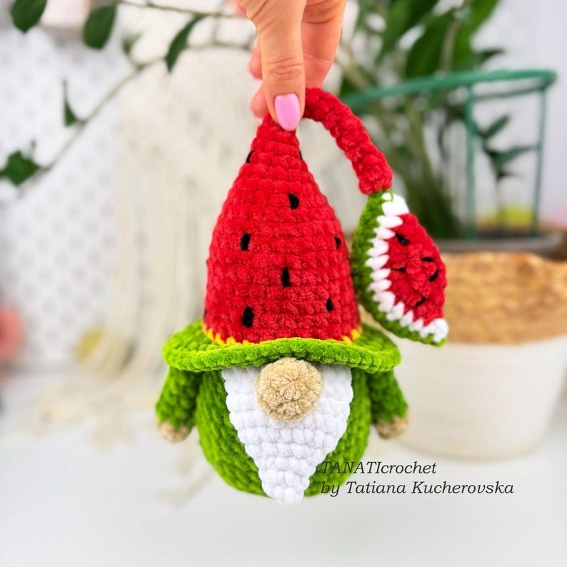 K&ouml;nnte beinhalten: Ein handgefertigter H&auml;kelzwerg mit Wassermelonen-Thema. Der Zwerg hat einen roten Hut mit schwarzen Samendetails, einen gr&uuml;nen K&ouml;rper und einen wei&szlig;en Bart. Ein kleines Wassermelonenscheibe ist befestigt. Der Zwerg wird von einer Hand gehalten.