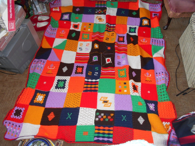 Może przedstawiać: Kolorowy, dziergany na szydełku koc z patchworkowym wzorem. Koc składa się z kwadrat&oacute;w w r&oacute;żnych kolorach, w tym czerwonym, pomarańczowym, ż&oacute;łtym, zielonym, niebieskim, fioletowym i białym.