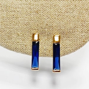 Peut inclure: Une paire de boucles d'oreilles à tige en or avec des pierres précieuses bleu foncé rectangulaires taillées en baguette. Chaque boucle d'oreille possède une monture en métal doré poli qui encadre la pierre bleue allongée.