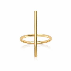 Peut inclure: Une bague en or composée d'un anneau lisse et poli surmonté d'une barre verticale unique fixée au centre. Le métal présente une finition brillante et réfléchissante, formant une silhouette géométrique épurée posée sur l'anneau circulaire.