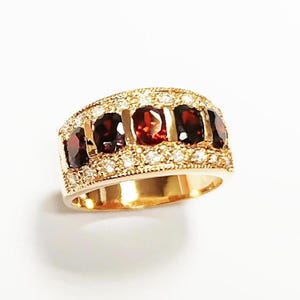 May include: A gold-toned ring featuring a row of oval-cut, deep red gemstones. The ring is encrusted with small, clear gemstones around the red stones and along the band, creating a sparkling effect. The ring is set against a white background.