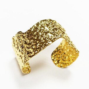 May include: A gold cuff bracelet with a textured, hammered finish. The open design features a wide band with an irregular, organic shape. The bracelet is set against a white background, highlighting its metallic sheen. The bracelet's dimensions are approximately 5 cm wide.