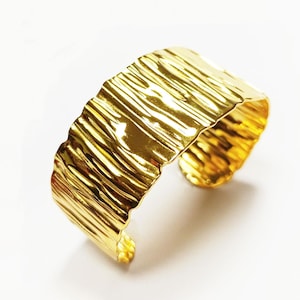 Pode incluir: Uma pulseira de punho dourada com um design texturizado e orgânico. A faixa larga apresenta uma superfície ondulada, criando um acessório único. O design aberto permite um uso fácil. Esta peça é uma adição ousada a qualquer coleção de joias.