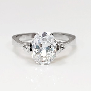 Peut inclure: Une bague en argent avec une grosse pierre précieuse claire de coupe ovale. La pierre précieuse est le point central, reflétant la lumière. La bague a un anneau simple avec des éléments décoratifs.