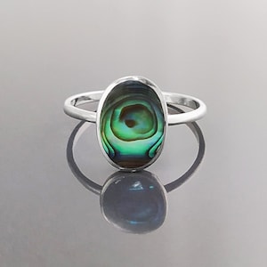 May include: A silver ring featuring an oval abalone shell inlay. The shell displays a green, blue, and brown iridescent pattern.