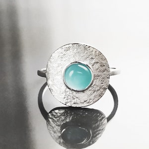 May include: A silver ring with a textured circular band and a light blue gemstone in the center.
