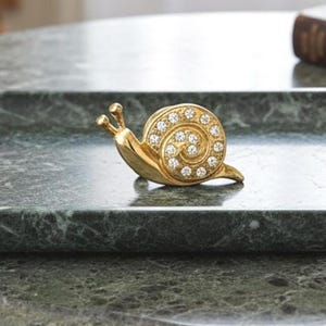 Puede incluir: Una figura de caracol en tono dorado con una concha en espiral adornada con brillantes piedras de estrás transparentes. El caracol tiene un cuerpo metálico liso y pulido con dos pequeñas antenas en la cabeza, un objeto decorativo detallado.