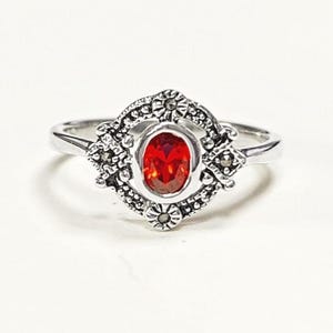 K&ouml;nnte beinhalten: Ein silberner Ring mit einem ovalen, roten Edelstein in der Mitte. Der Edelstein ist von einer dekorativen, floral inspirierten Fassung umgeben. Der Ring hat ein schlankes, poliertes Band und ein elegantes, vintage-inspiriertes Design.