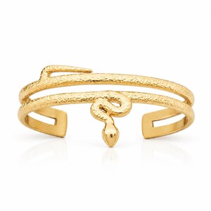 May include: A gold-toned cuff bracelet featuring a double-band design with a textured surface. The bracelet is shaped like a coiled snake, with the head of the snake positioned at the center of the front opening, creating a wrap-around effect.