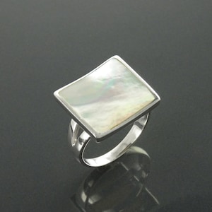 Puede incluir: Un anillo de plata con una gran piedra rectangular iridiscente de nácar. El anillo tiene un diseño simple y elegante con un acabado pulido. El nácar muestra una gama de colores, incluyendo blanco, plata y toques de verde.