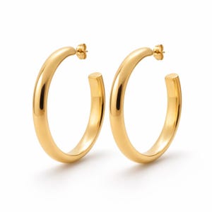 Peut inclure: Une paire de boucles d'oreilles créoles dorées. Les boucles d'oreilles ont une surface lisse et polie et un design circulaire classique. Les tiges sont visibles. Elles sont sur fond blanc.
