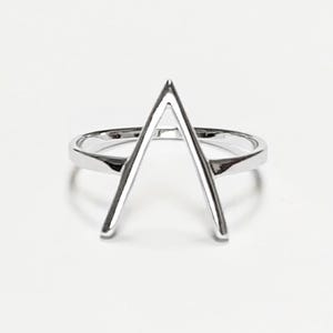 Könnte beinhalten: Ein silberner Ring mit minimalistischem Design. Der Ring hat eine dreieckige Form in der Mitte, wobei der Scheitel nach oben zeigt. Das Band ist glatt und poliert, und der Ring ist vor einem weißen Hintergrund platziert.