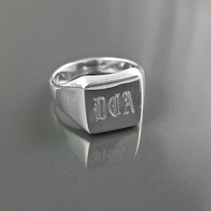 Peut inclure: Une bague chevalière en argent poli avec une face rectangulaire. La face est gravée des lettres "DDA" dans une police ornée. La bague est très brillante et reflète la lumière.