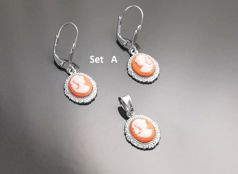 May include: Set A of silver earrings and a pendant featuring a cameo design with a white profile on an orange background. The earrings have a lever back closure.