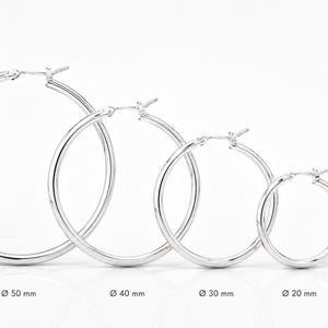 Puede incluir: Cinco pendientes de aro plateados dispuestos en fila de mayor a menor. El grosor del alambre es de 2 mm. Los diámetros se indican como 50 mm, 40 mm, 30 mm, 20 mm y 14 mm. Cada pendiente presenta un diseño circular simple con cierre articulado.