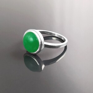 May include: A silver ring featuring a large, round, bright green gemstone. The gemstone is set in a bezel setting, and the ring band is a simple, polished silver. The ring is displayed on a reflective surface.