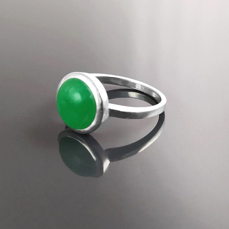 May include: A silver ring featuring a large, round, bright green gemstone. The gemstone is set in a bezel setting, and the ring band is a simple, polished silver. The ring is displayed on a reflective surface.