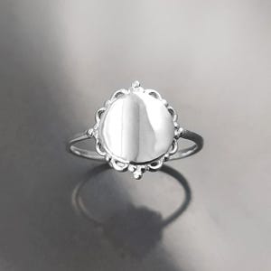 Op de afbeelding: Een zilveren ring met een ronde, lege middenstuk en een decoratieve geschulpte rand. De ring heeft een dunne band en een symmetrisch ontwerp. Gepresenteerd op een reflecterend oppervlak.