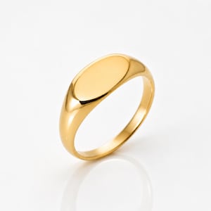 May include: A polished gold signet ring featuring a smooth, oval-shaped flat top surface. The band is solid and tapers slightly toward the bottom, showcasing a reflective metallic finish that catches the light across its curved, seamless exterior.