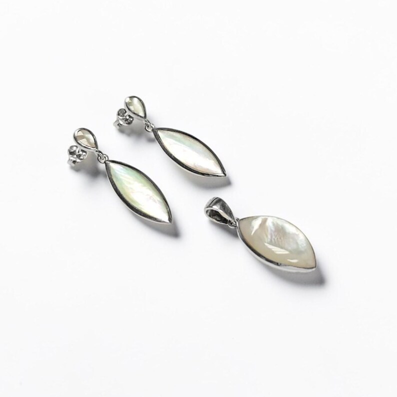 May include: A pair of silver drop earrings and a matching pendant, each featuring an elongated marquise-shaped inlay of iridescent white mother-of-pearl. The metal settings are polished silver, with the earrings using a post-back closure.